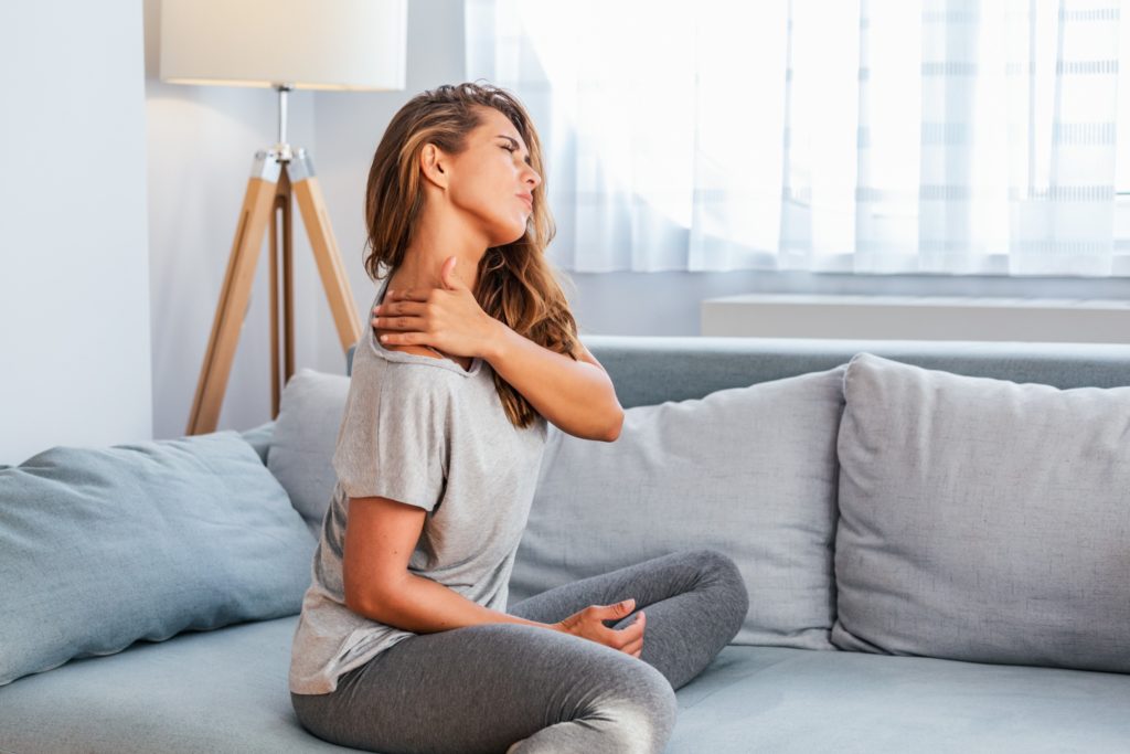 Blonde woman wearing athletic clothes sitting on a gray couch with her neck stretch and her left hand rubbing her right shoulder
