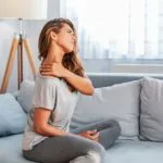 Blonde woman wearing athletic clothes sitting on a gray couch with her neck stretch and her left hand rubbing her right shoulder