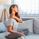 Blonde woman wearing athletic clothes sitting on a gray couch with her neck stretch and her left hand rubbing her right shoulder