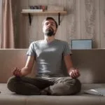 Image of a white man with dark hair and a beard sitting on a couch with a phone and laptop by his side. His eyes are closed and he is sitting cross-legged appearing to meditate.