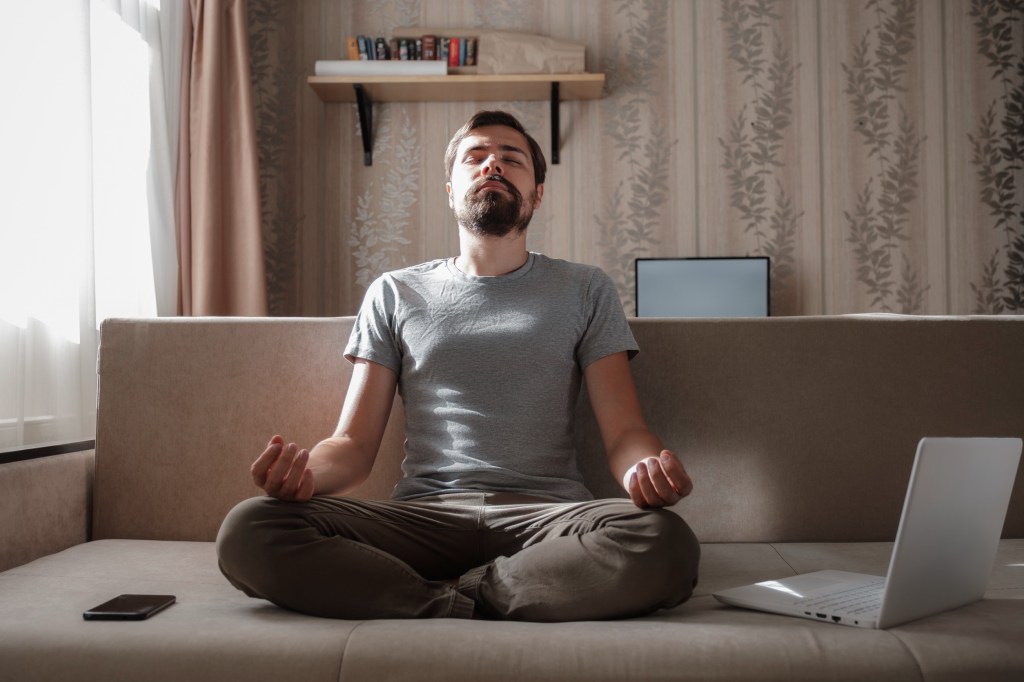 Image of a white man with dark hair and a beard sitting on a couch with a phone and laptop by his side. His eyes are closed and he is sitting cross-legged appearing to meditate.